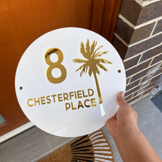 A circular white and gold coastal house sign that features the house number, street name and a detailed palm tree . Made from UV stable acrylic suitable for external use.